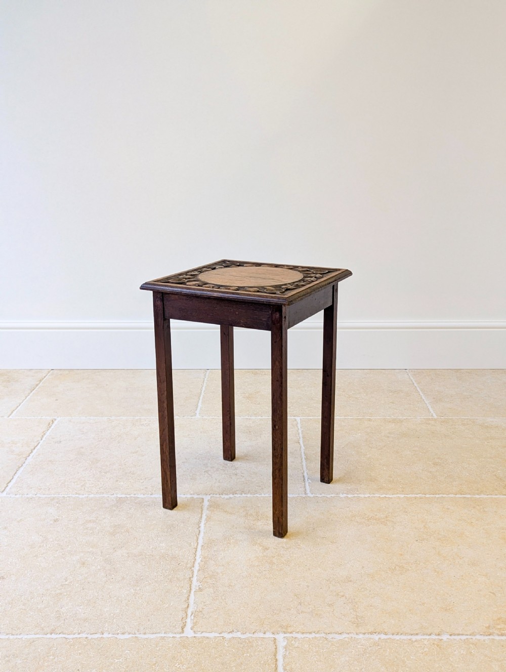 A square dark oak occasional table featuring a relief-carved floral border around a central circular panel, supported by four straight square legs. View X