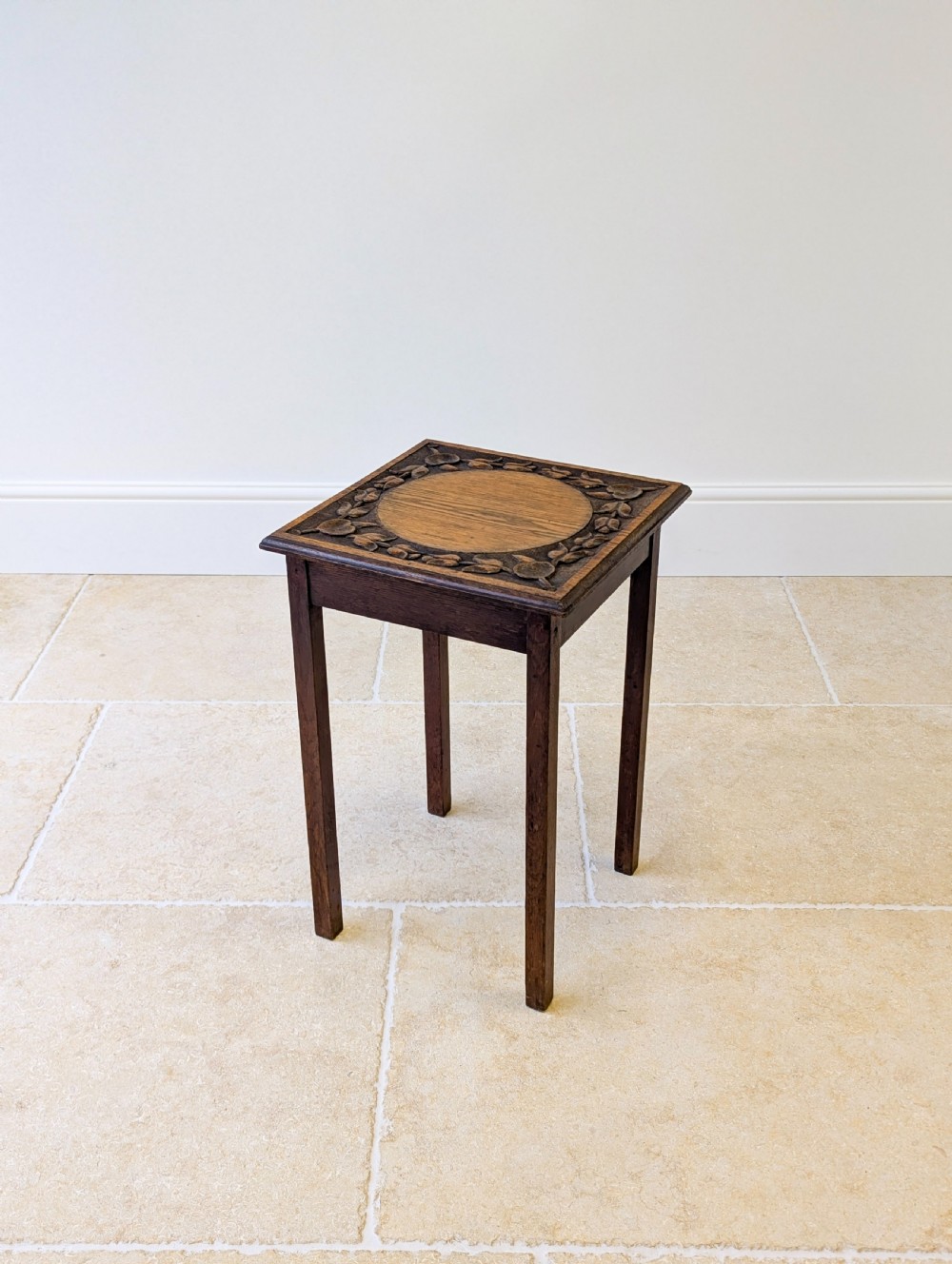 A square dark oak occasional table featuring a relief-carved floral border around a central circular panel, supported by four straight square legs. View XII