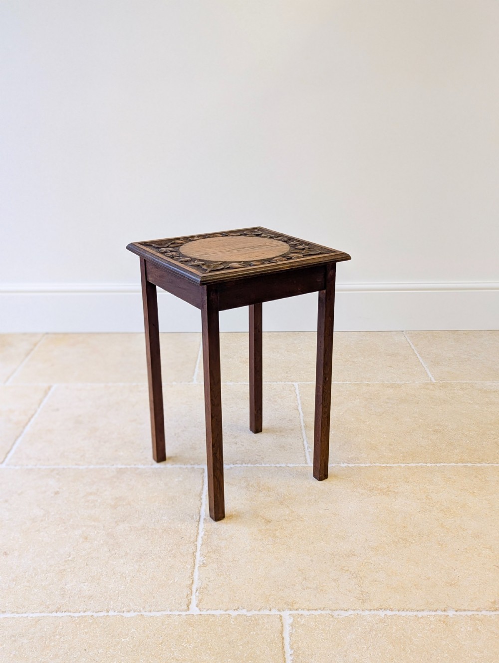 A square dark oak occasional table featuring a relief-carved floral border around a central circular panel, supported by four straight square legs. View XV