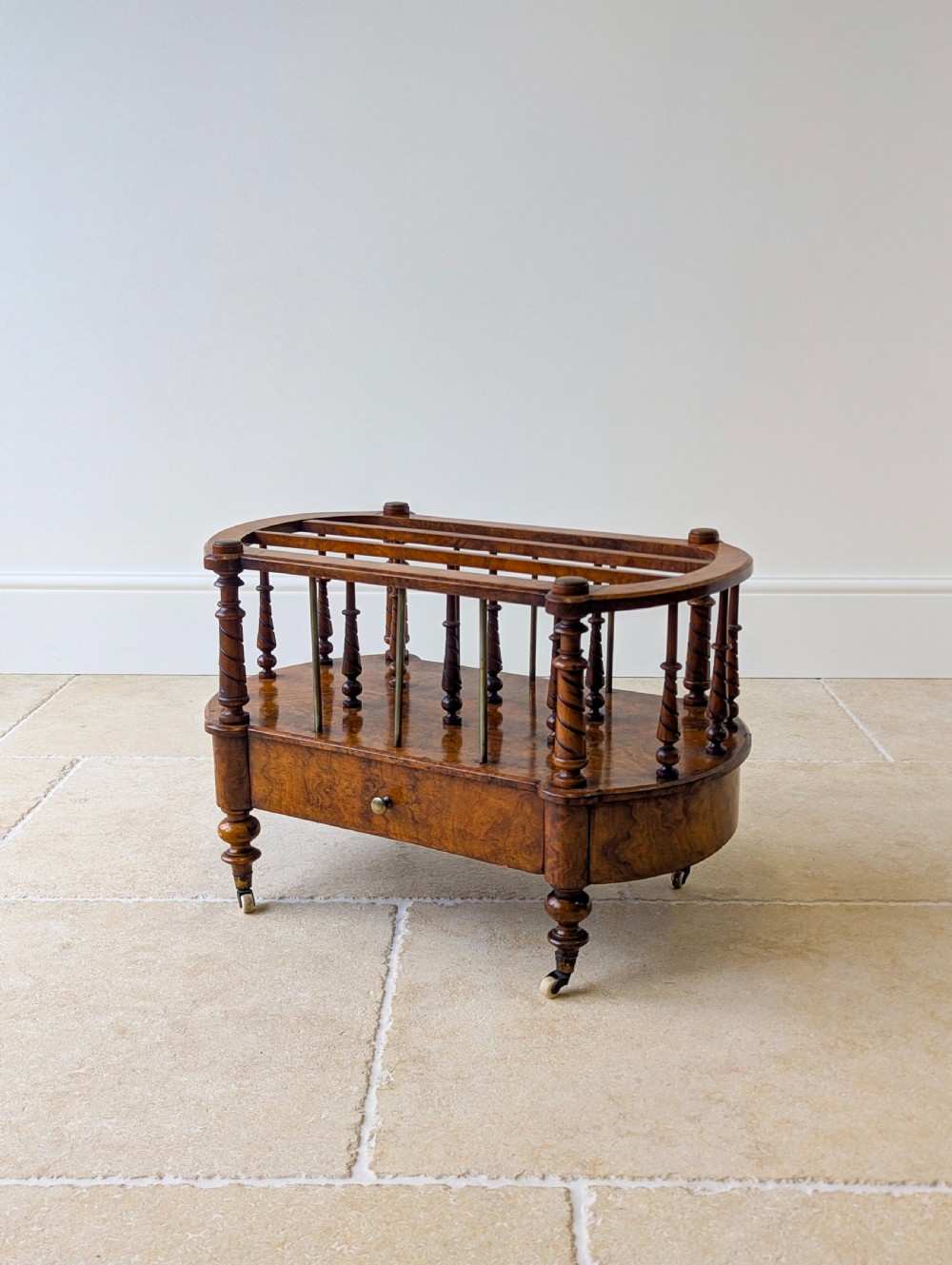 A Victorian burr walnut canterbury featuring turned spiral columns, brass division rods, a base drawer, and ceramic castors.