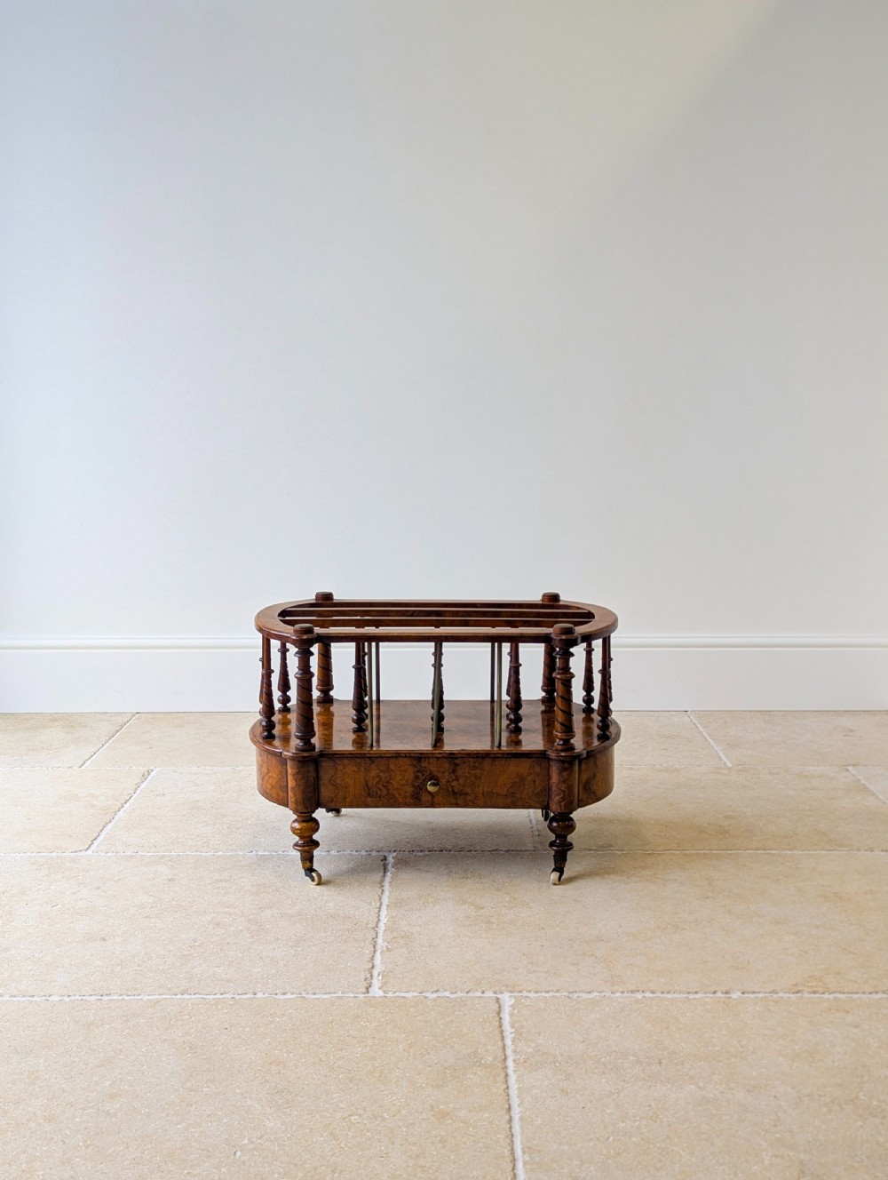 A Victorian burr walnut canterbury featuring turned spiral columns, brass division rods, a base drawer, and ceramic castors. View V
