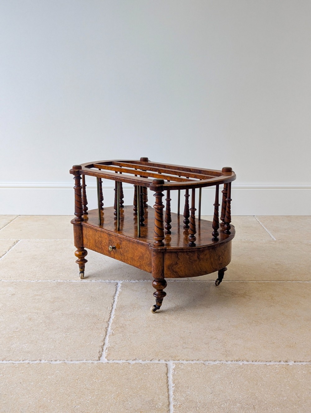 A Victorian burr walnut canterbury featuring turned spiral columns, brass division rods, a base drawer, and ceramic castors. View XV