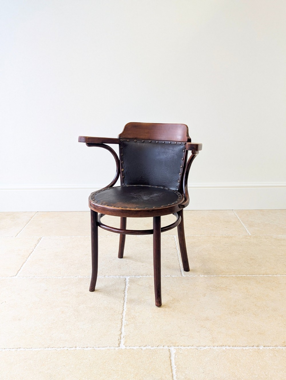 An antique Edwardian bentwood armchair featuring dark brown steam-bent beech wood, sweeping armrests, and a black leather studded seat and backrest.