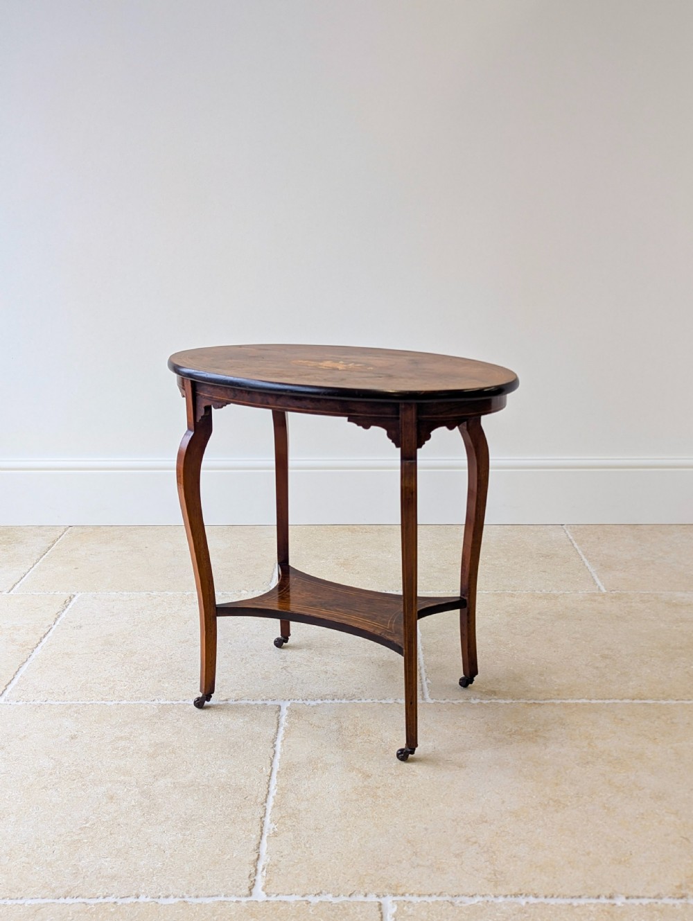 An Edwardian rosewood oval occasional table with marquetry inlay on the tabletop, slender cabriole legs, a shaped lower shelf, and brass castors.