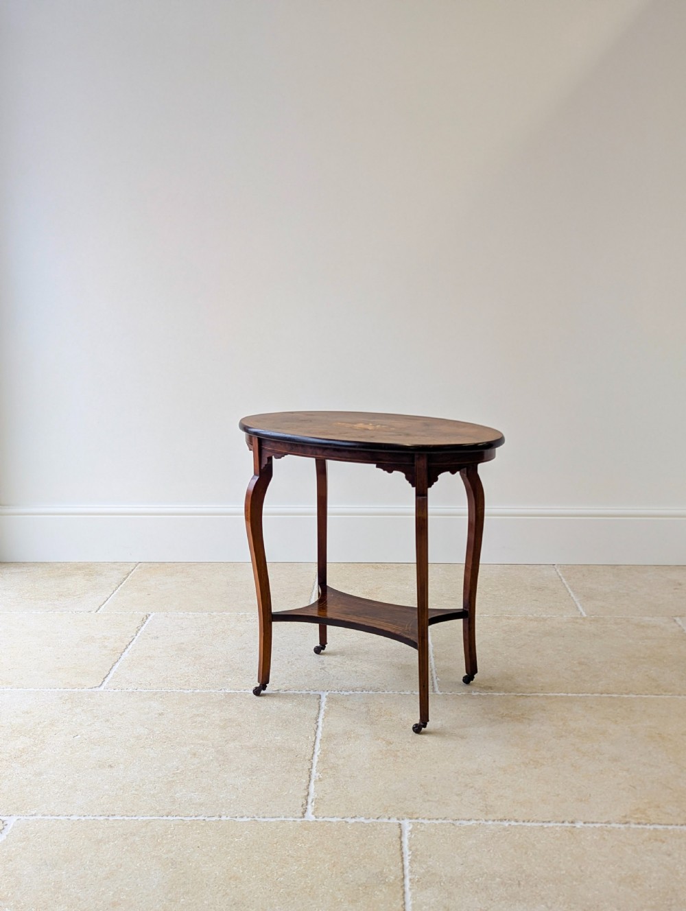An Edwardian rosewood oval occasional table with marquetry inlay on the tabletop, slender cabriole legs, a shaped lower shelf, and brass castors. View V