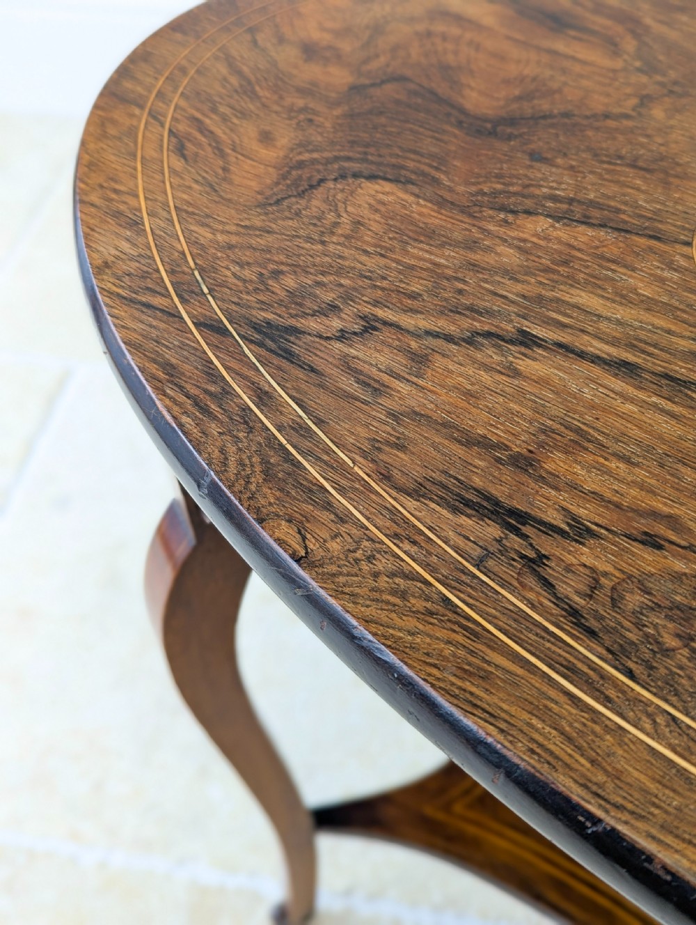 An Edwardian rosewood oval occasional table with marquetry inlay on the tabletop, slender cabriole legs, a shaped lower shelf, and brass castors. View VI