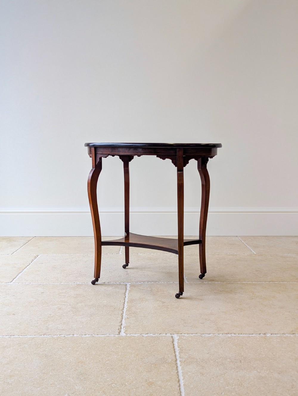 An Edwardian rosewood oval occasional table with marquetry inlay on the tabletop, slender cabriole legs, a shaped lower shelf, and brass castors. View IX