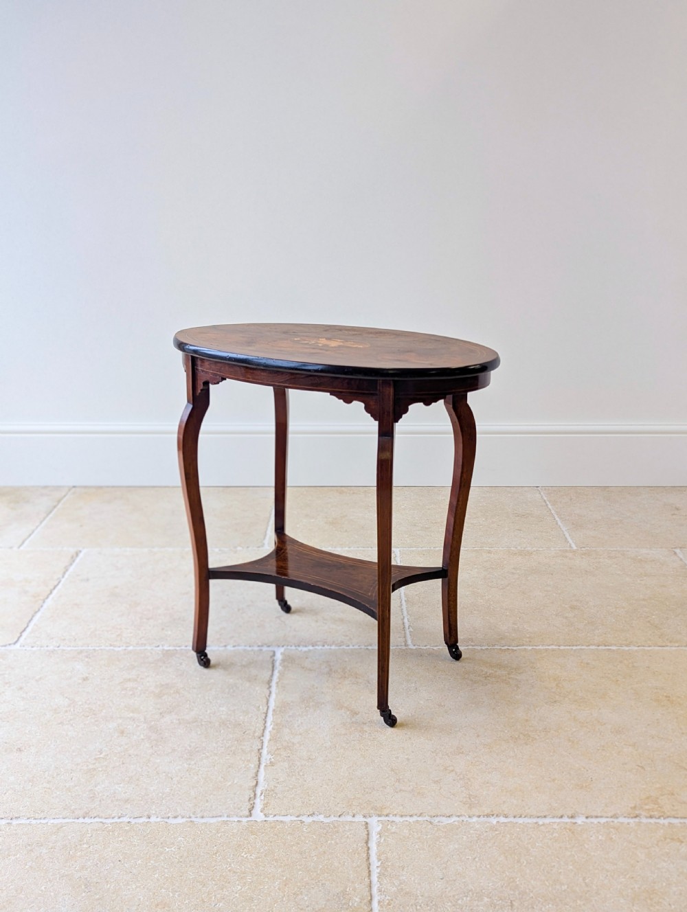 An Edwardian rosewood oval occasional table with marquetry inlay on the tabletop, slender cabriole legs, a shaped lower shelf, and brass castors. View XIII