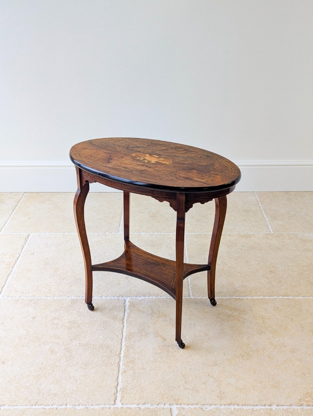 An Edwardian rosewood oval occasional table with marquetry inlay on the tabletop, slender cabriole legs, a shaped lower shelf, and brass castors. View XV
