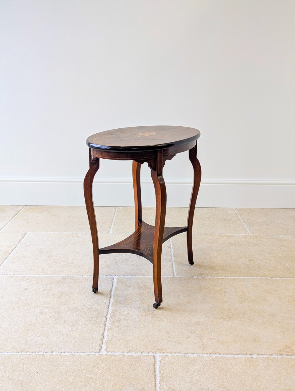 An Edwardian rosewood oval occasional table with marquetry inlay on the tabletop, slender cabriole legs, a shaped lower shelf, and brass castors. View XVI