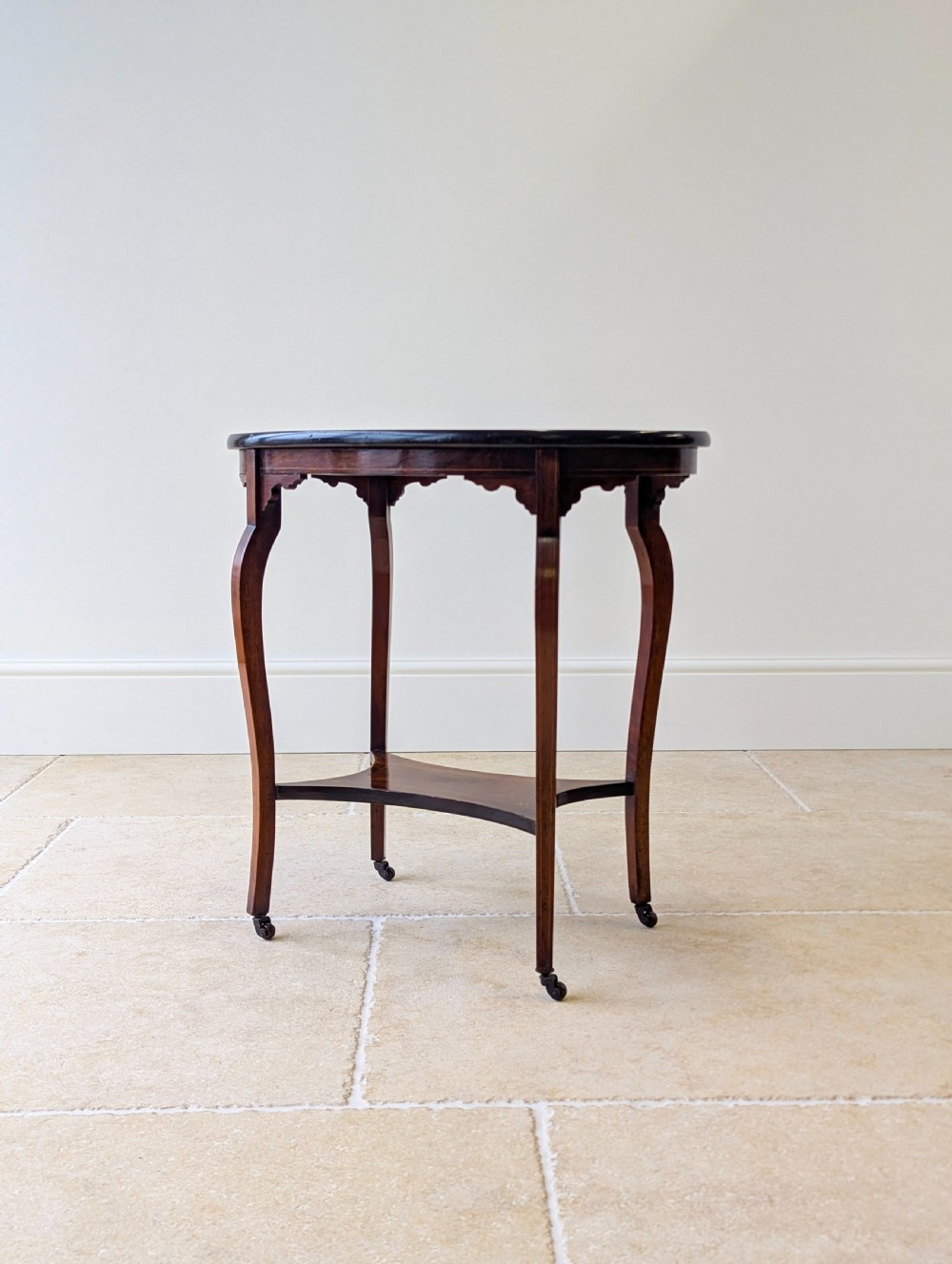 An Edwardian rosewood oval occasional table with marquetry inlay on the tabletop, slender cabriole legs, a shaped lower shelf, and brass castors. View XVII