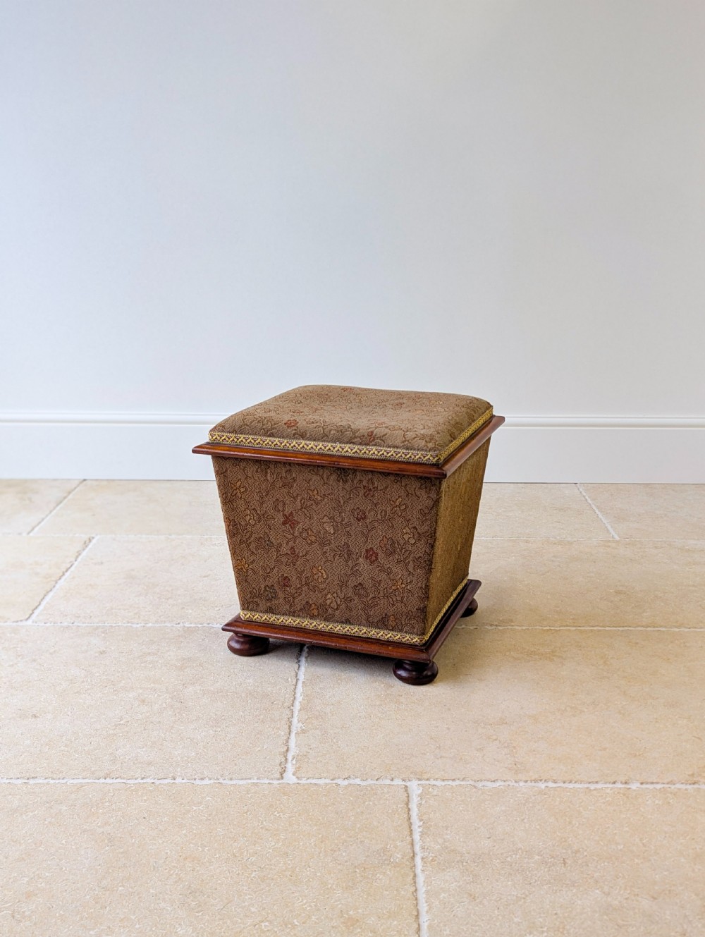 A Victorian upholstered mahogany storage ottoman featuring a tapered body, brown patterned fabric, braided trim, and turned bun feet.
