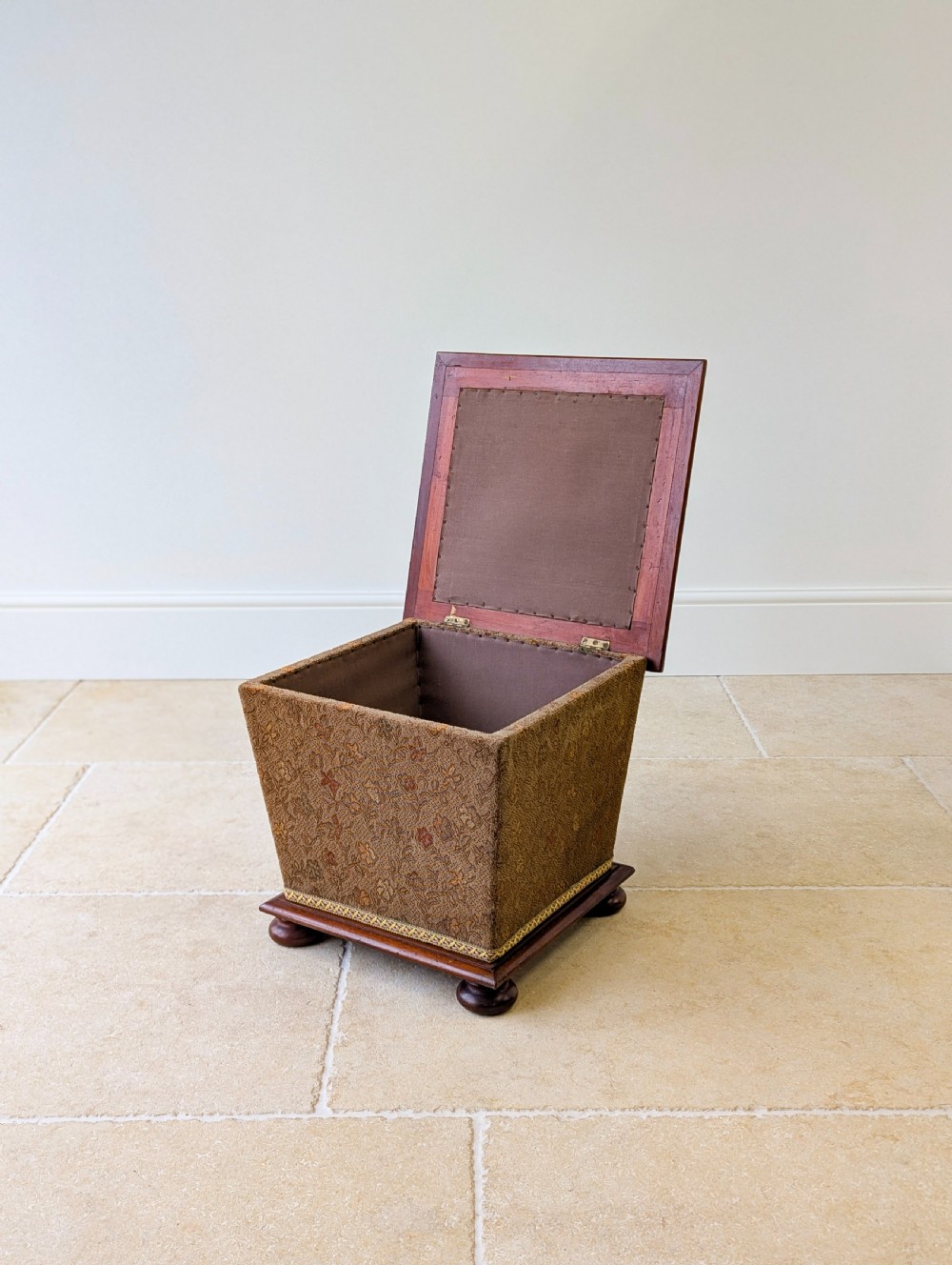 A Victorian upholstered mahogany storage ottoman featuring a tapered body, brown patterned fabric, braided trim, and turned bun feet. View II