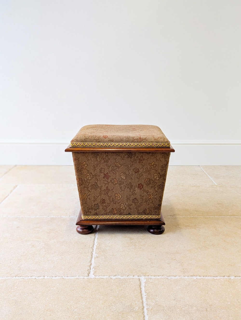 A Victorian upholstered mahogany storage ottoman featuring a tapered body, brown patterned fabric, braided trim, and turned bun feet. View III