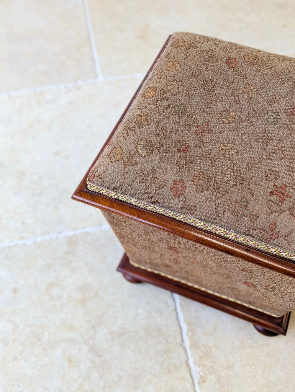 A Victorian upholstered mahogany storage ottoman featuring a tapered body, brown patterned fabric, braided trim, and turned bun feet. View IV
