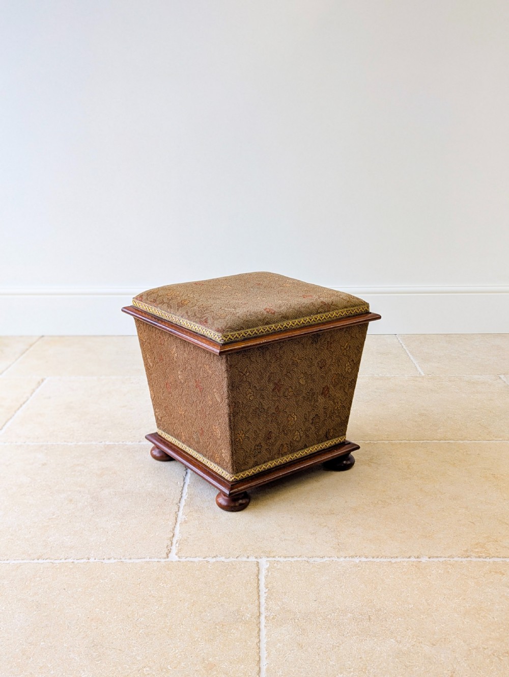 A Victorian upholstered mahogany storage ottoman featuring a tapered body, brown patterned fabric, braided trim, and turned bun feet. View V