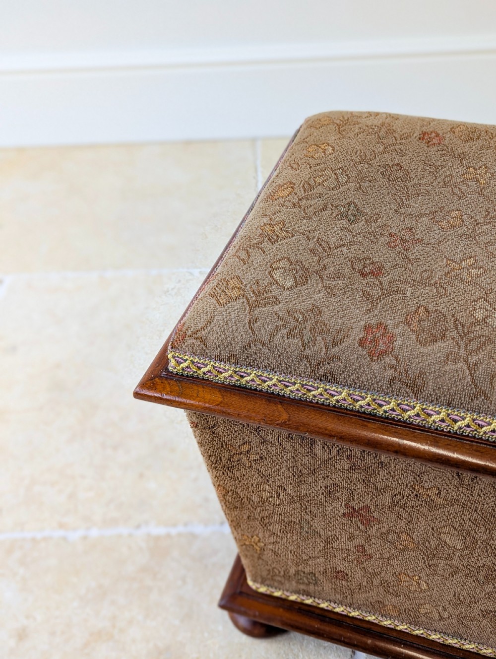 A Victorian upholstered mahogany storage ottoman featuring a tapered body, brown patterned fabric, braided trim, and turned bun feet. View VI