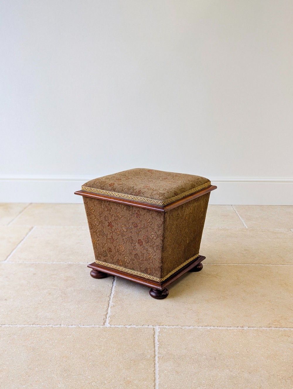 A Victorian upholstered mahogany storage ottoman featuring a tapered body, brown patterned fabric, braided trim, and turned bun feet. View VII
