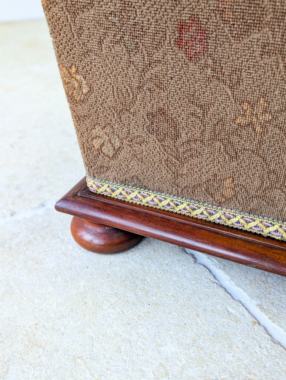 A Victorian upholstered mahogany storage ottoman featuring a tapered body, brown patterned fabric, braided trim, and turned bun feet. View VIII