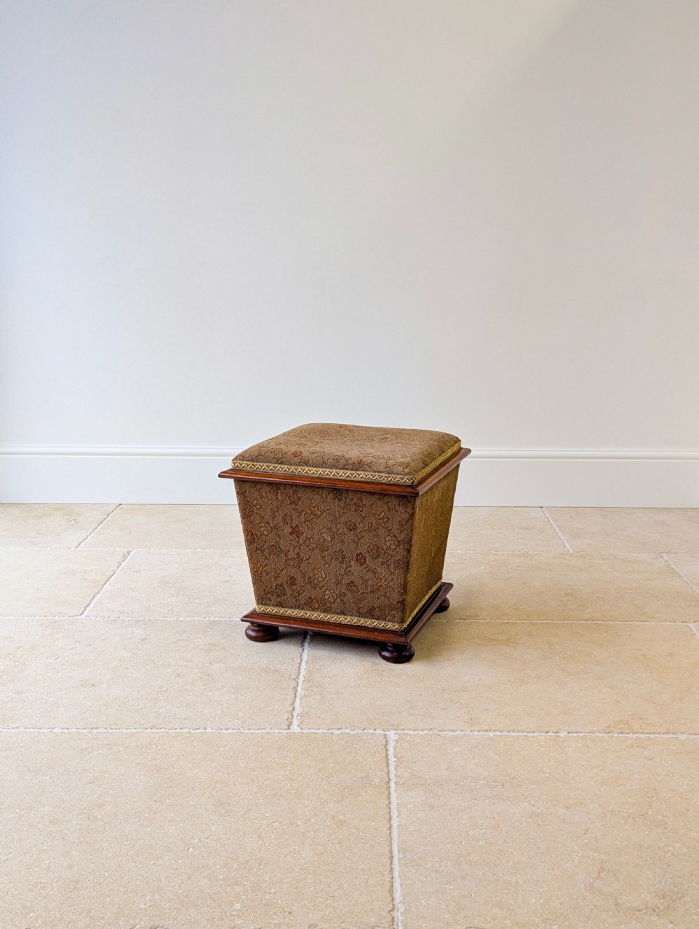 A Victorian upholstered mahogany storage ottoman featuring a tapered body, brown patterned fabric, braided trim, and turned bun feet. View IX