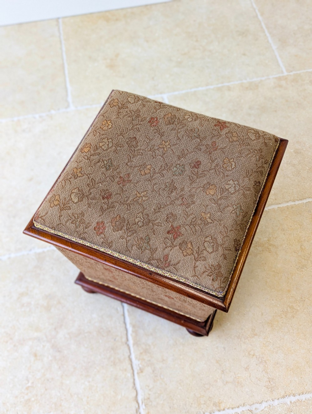A Victorian upholstered mahogany storage ottoman featuring a tapered body, brown patterned fabric, braided trim, and turned bun feet. View X