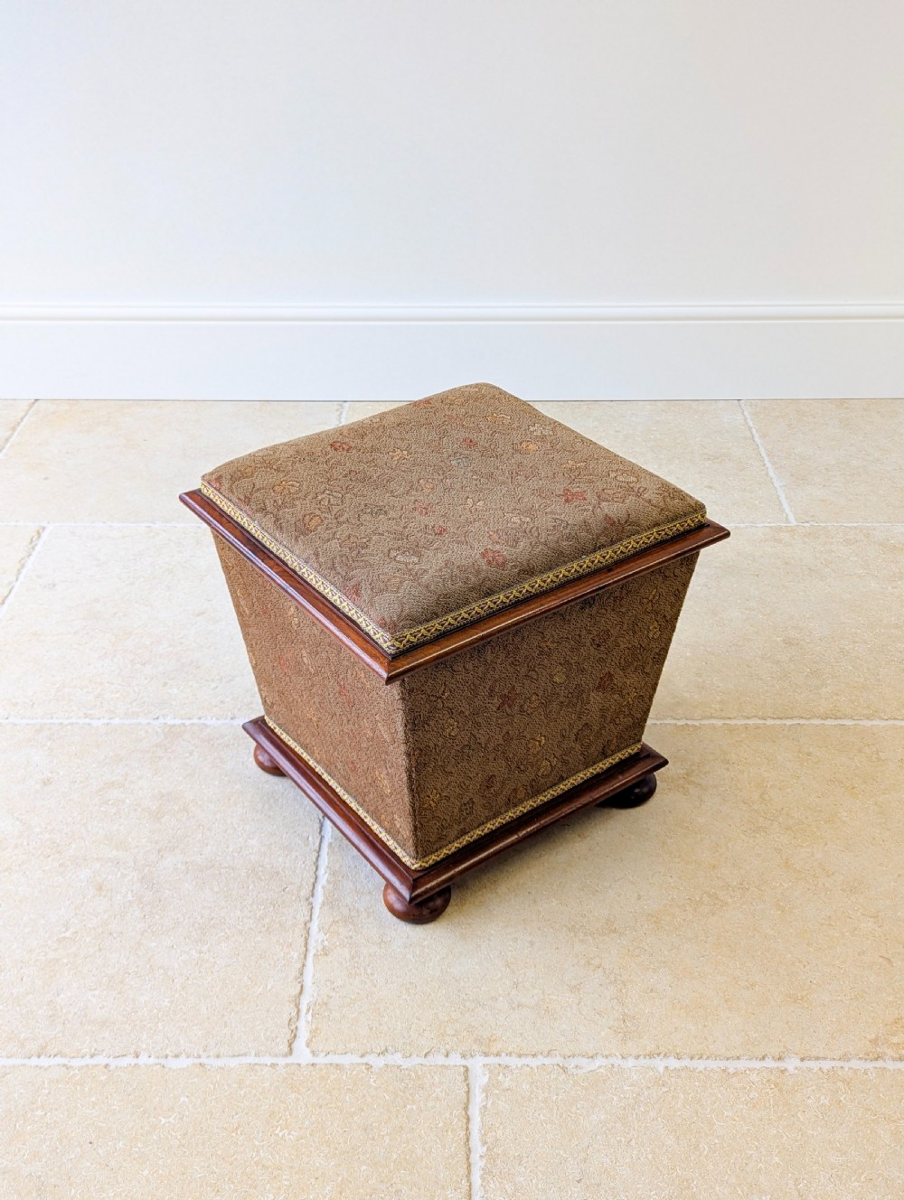 A Victorian upholstered mahogany storage ottoman featuring a tapered body, brown patterned fabric, braided trim, and turned bun feet. View XI