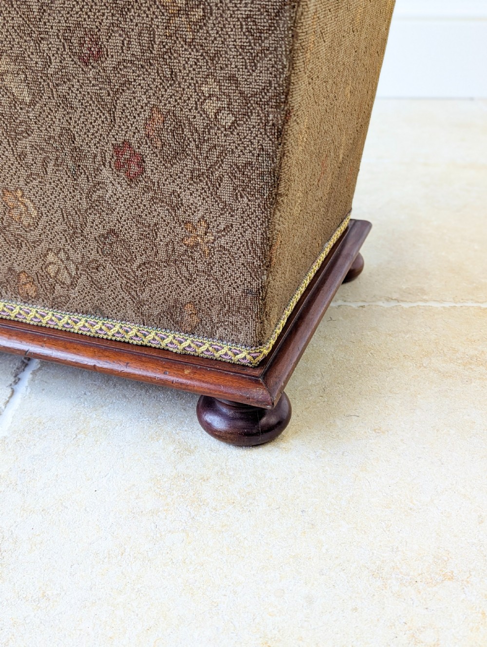 A Victorian upholstered mahogany storage ottoman featuring a tapered body, brown patterned fabric, braided trim, and turned bun feet. View XII
