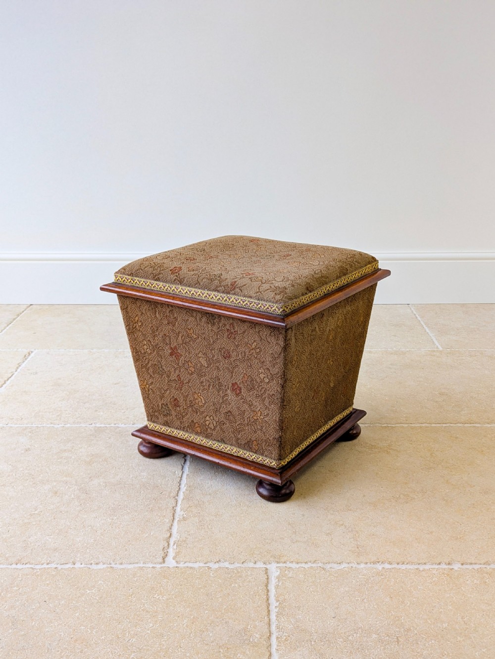 A Victorian upholstered mahogany storage ottoman featuring a tapered body, brown patterned fabric, braided trim, and turned bun feet. View XIII
