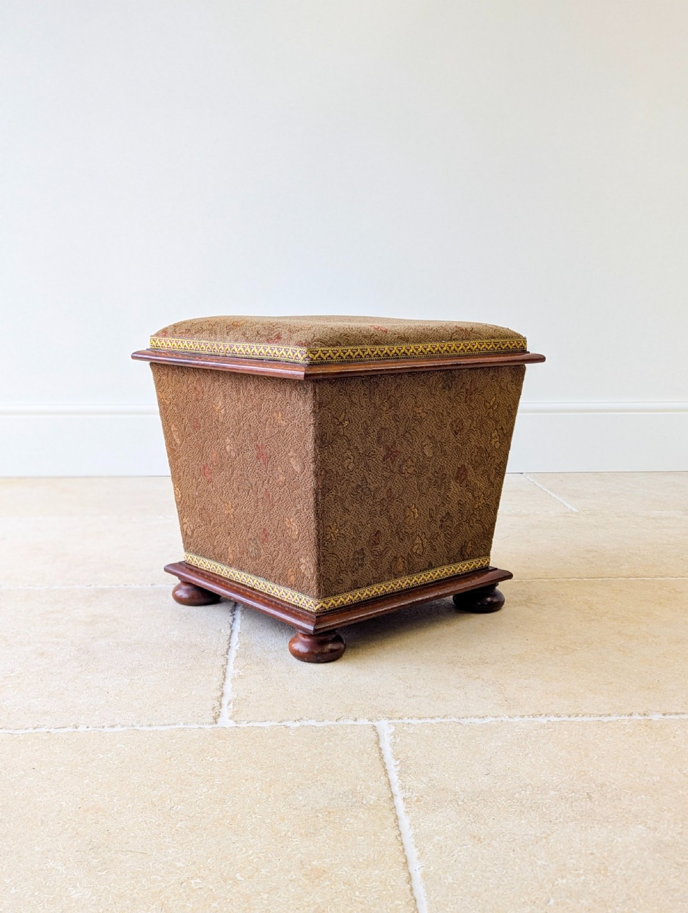 A Victorian upholstered mahogany storage ottoman featuring a tapered body, brown patterned fabric, braided trim, and turned bun feet. View XV