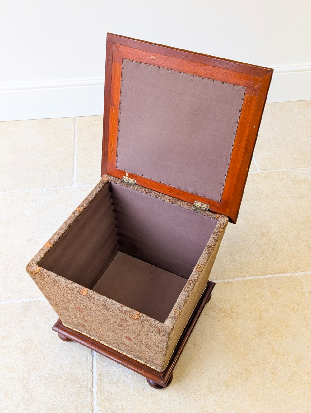 A Victorian upholstered mahogany storage ottoman featuring a tapered body, brown patterned fabric, braided trim, and turned bun feet. View XVI