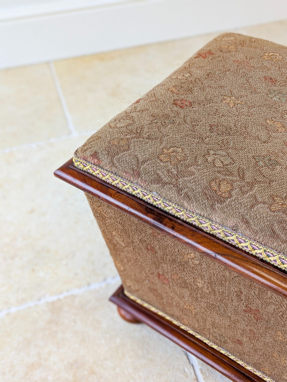 A Victorian upholstered mahogany storage ottoman featuring a tapered body, brown patterned fabric, braided trim, and turned bun feet. View XVII