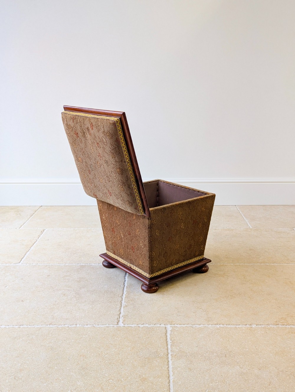 A Victorian upholstered mahogany storage ottoman featuring a tapered body, brown patterned fabric, braided trim, and turned bun feet. View XVIII