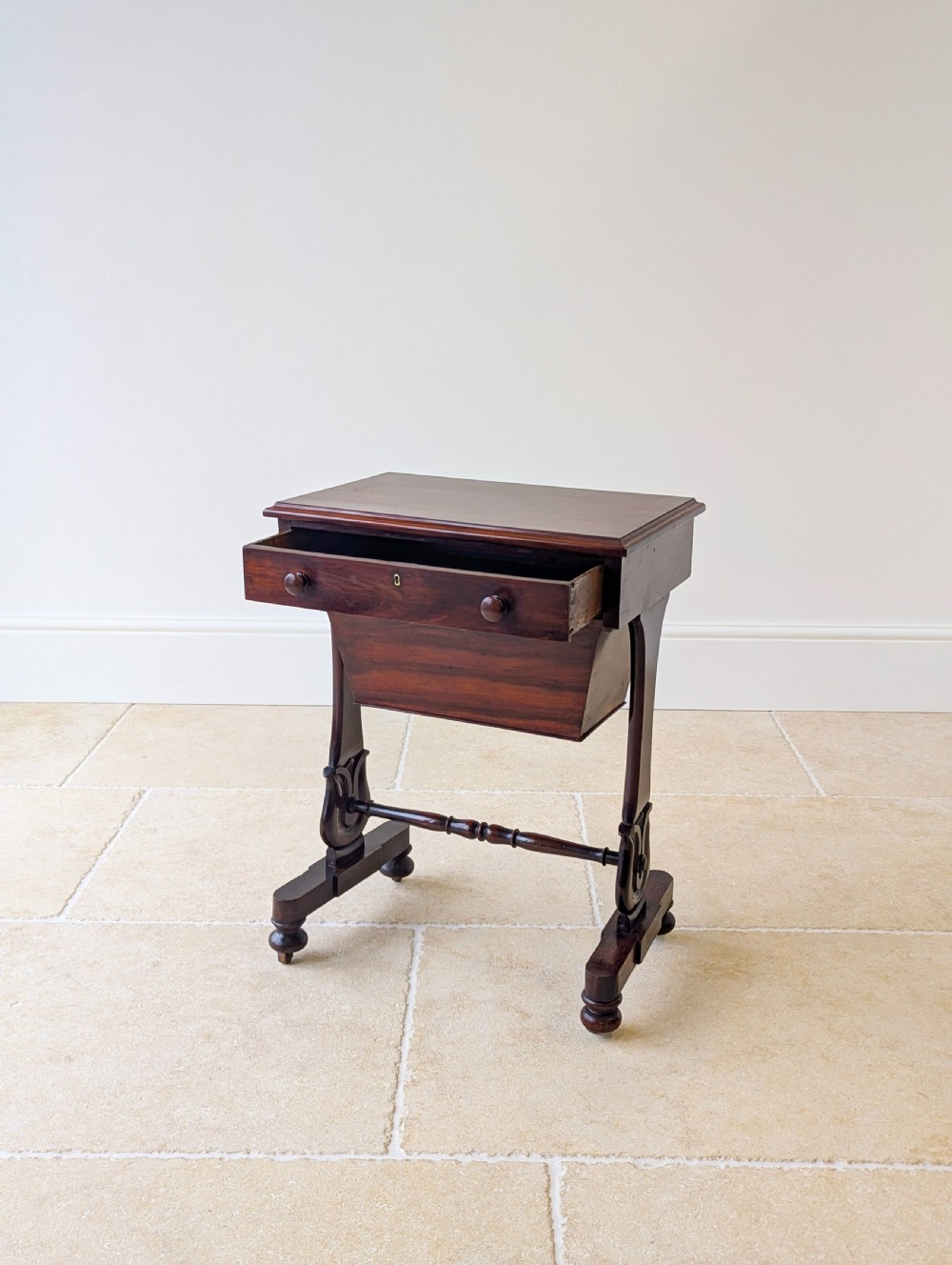 A late 19th-century Victorian mahogany sewing table featuring a top drawer with circular turned wooden handles over a deep tapered work bin, raised on scrolled lyre side supports, a turned stretcher, and castors. View IV