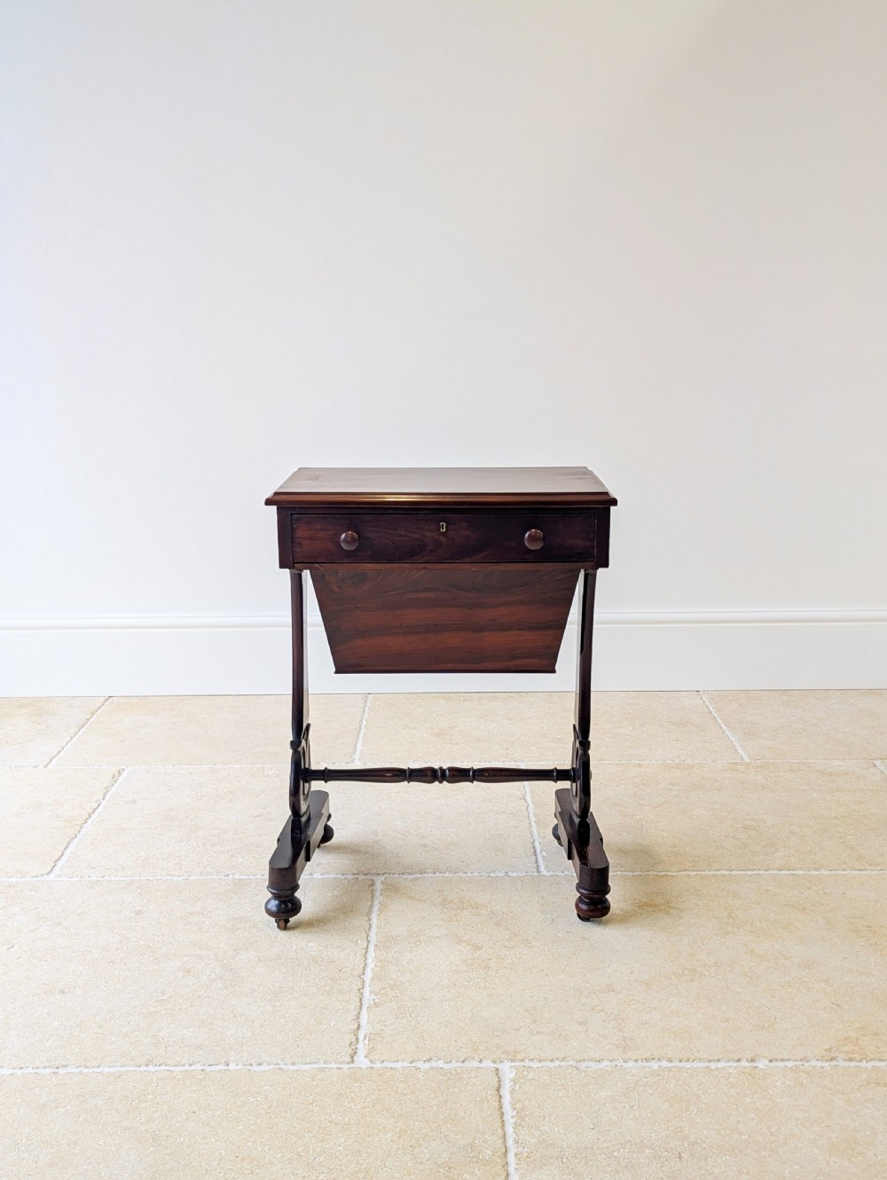 A late 19th-century Victorian mahogany sewing table featuring a top drawer with circular turned wooden handles over a deep tapered work bin, raised on scrolled lyre side supports, a turned stretcher, and castors. View VI