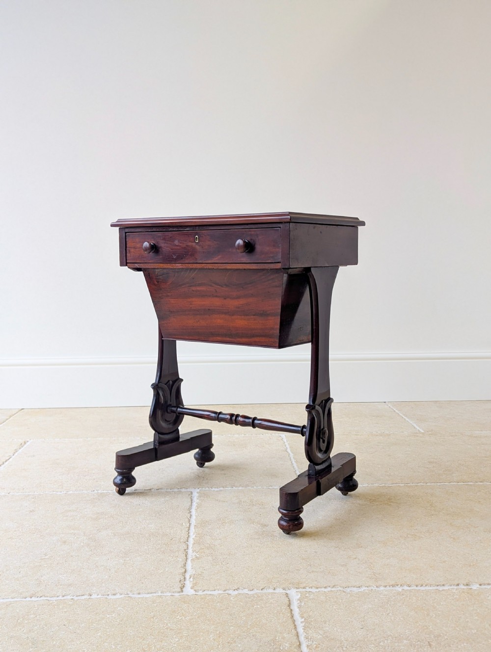 A late 19th-century Victorian mahogany sewing table featuring a top drawer with circular turned wooden handles over a deep tapered work bin, raised on scrolled lyre side supports, a turned stretcher, and castors. View XII