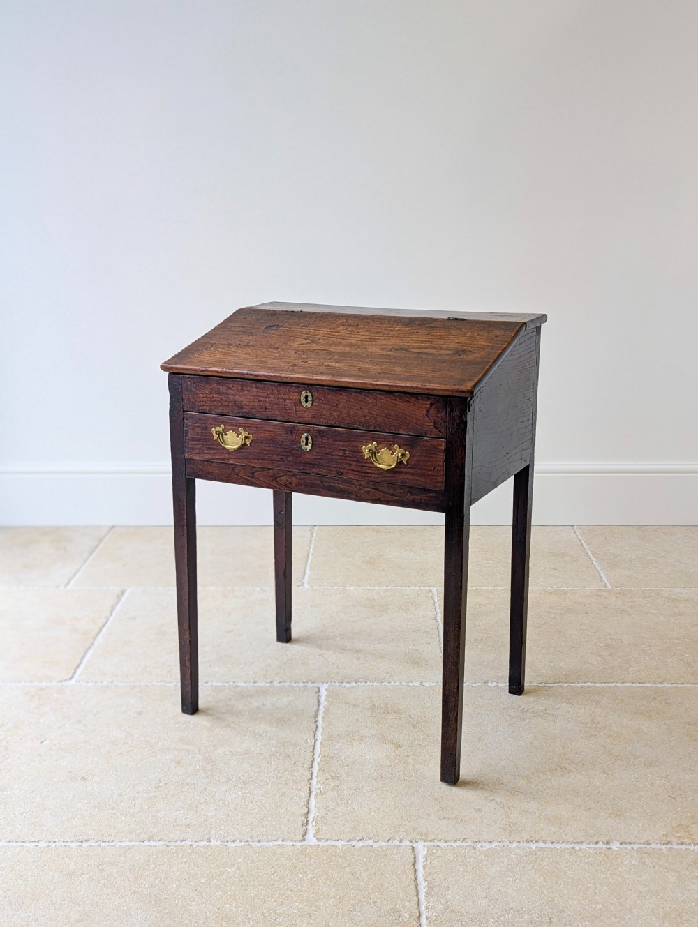 A Georgian dark elm clerks desk featuring a sloped writing surface, brass escutcheons, brass handles, and square legs.