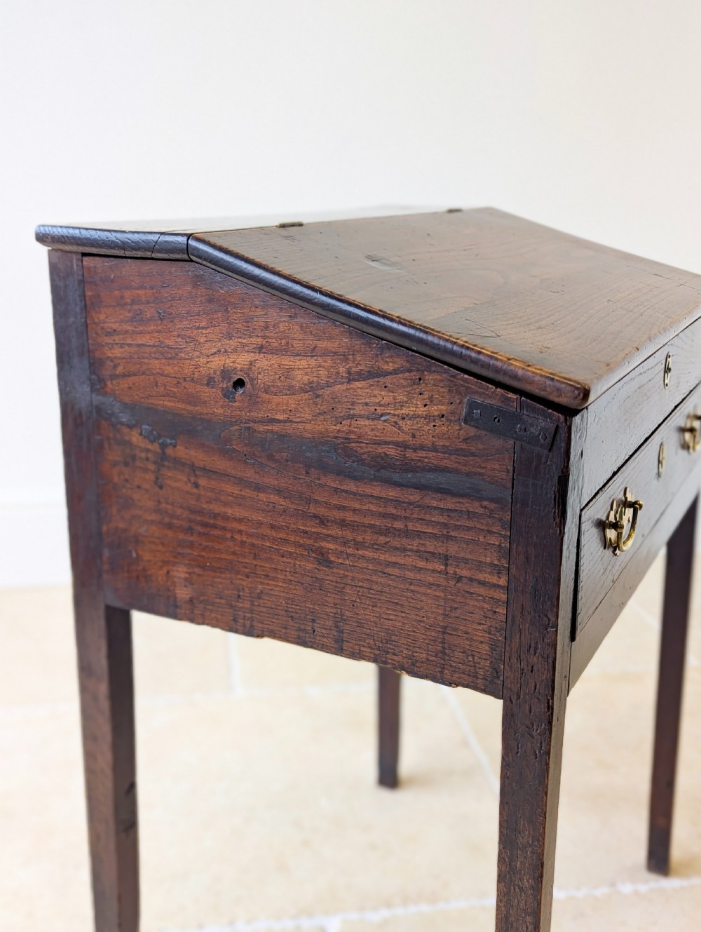 A Georgian dark elm clerks desk featuring a sloped writing surface, brass escutcheons, brass handles, and square legs. View XV