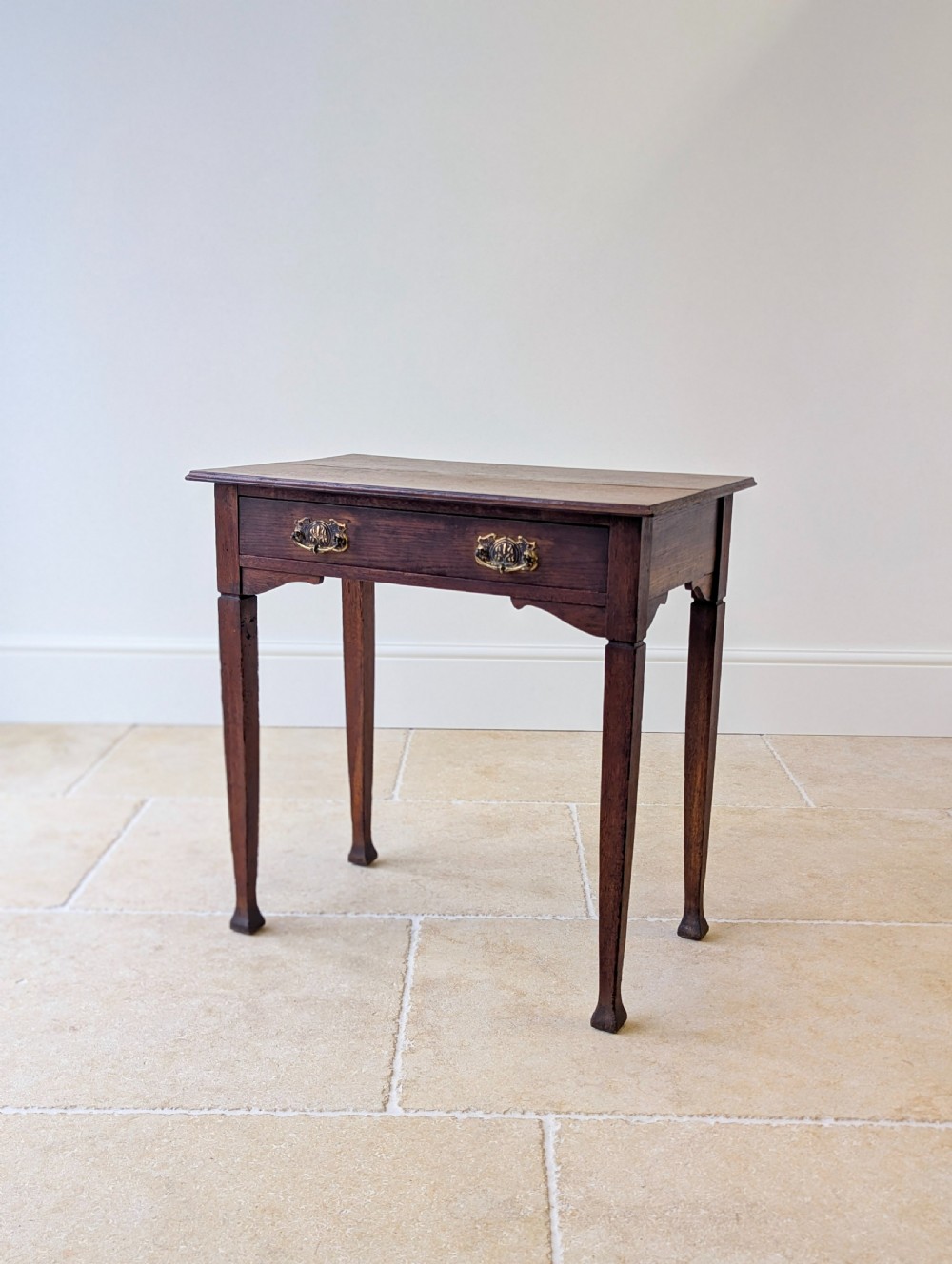 An antique dark oak side table featuring a single drawer with brass drop handles, shaped corner aprons, and slender tapered legs.
