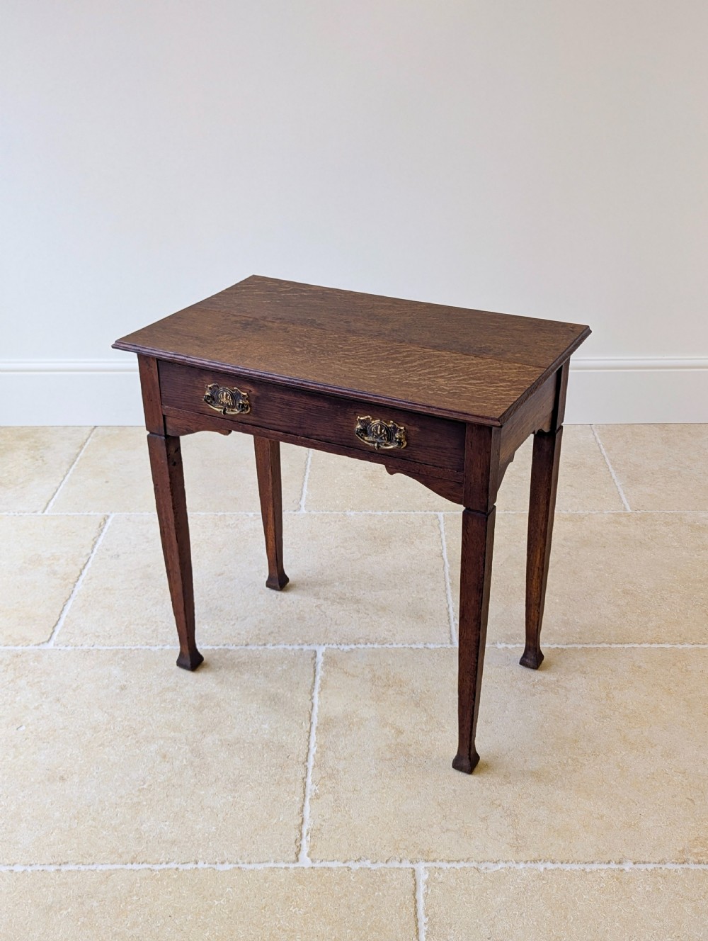 An antique dark oak side table featuring a single drawer with brass drop handles, shaped corner aprons, and slender tapered legs. View X