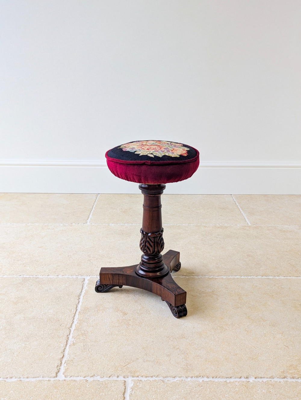 A William IV rosewood adjustable piano stool featuring a circular floral needlepoint seat with burgundy velvet trim, supported by a carved pedestal and triform base.