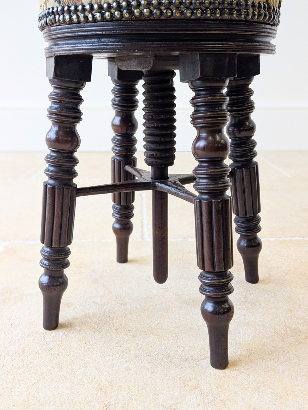 A Victorian dark mahogany adjustable piano stool featuring four turned legs, a threaded centre spindle, and a circular patterned fabric seat trimmed with brass studs. View IV