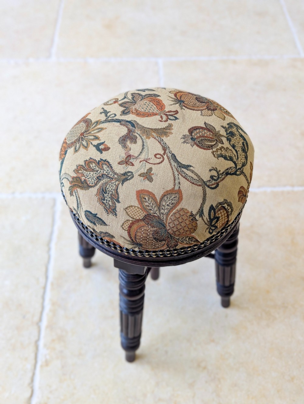 A Victorian dark mahogany adjustable piano stool featuring four turned legs, a threaded centre spindle, and a circular patterned fabric seat trimmed with brass studs. View VI