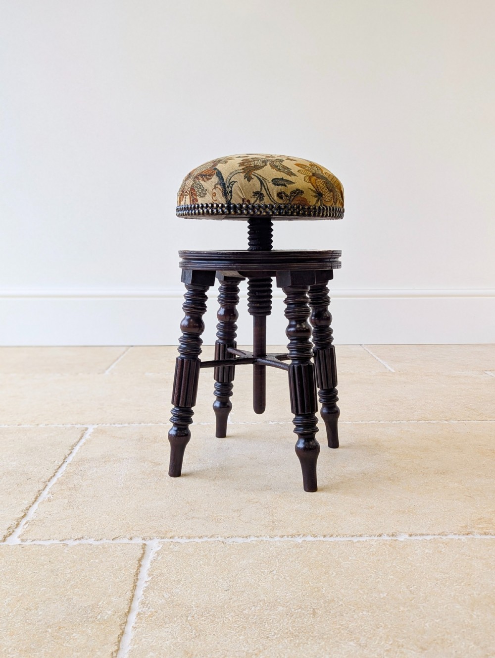 A Victorian dark mahogany adjustable piano stool featuring four turned legs, a threaded centre spindle, and a circular patterned fabric seat trimmed with brass studs. View VII