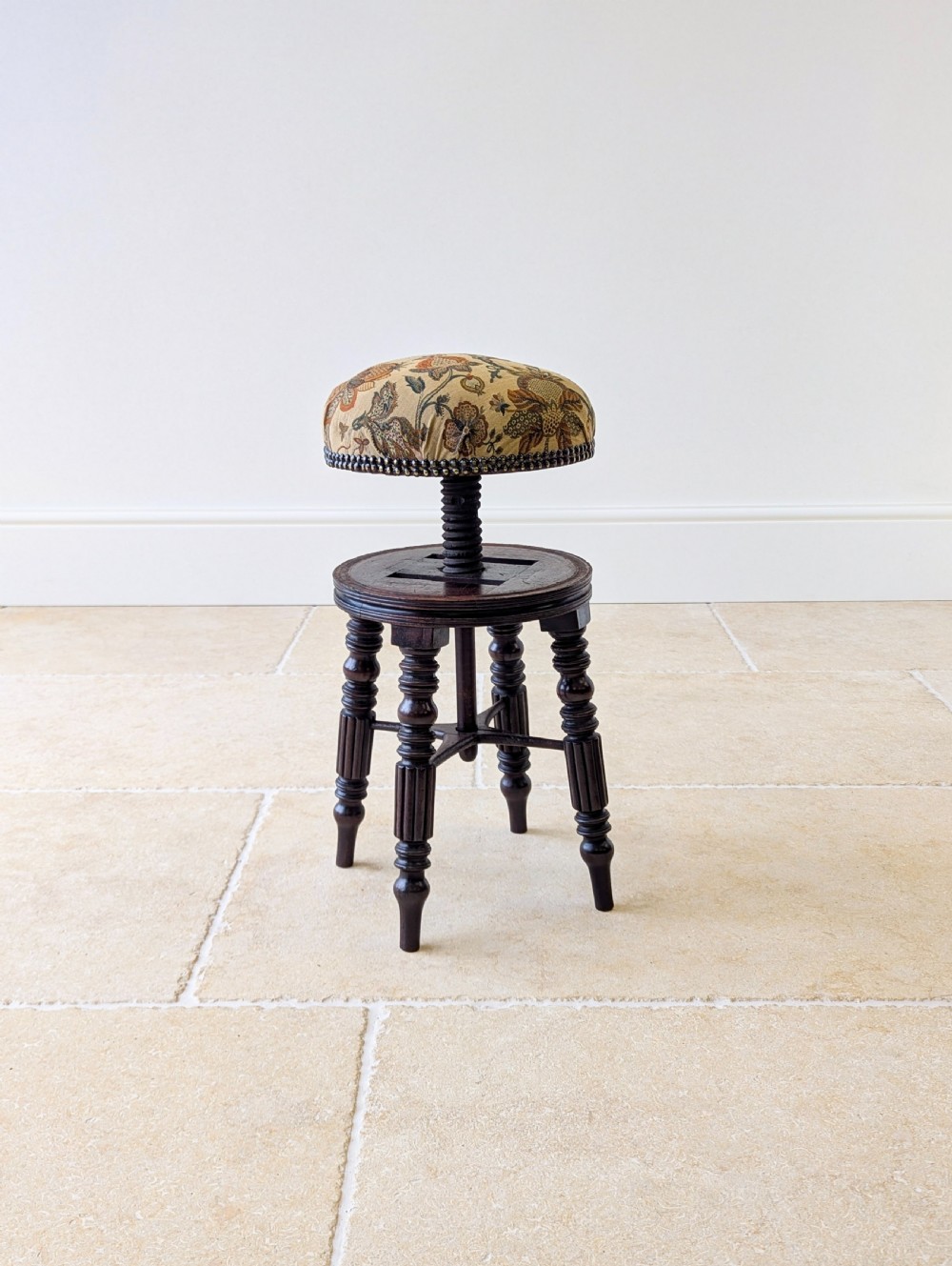 A Victorian dark mahogany adjustable piano stool featuring four turned legs, a threaded centre spindle, and a circular patterned fabric seat trimmed with brass studs. View X