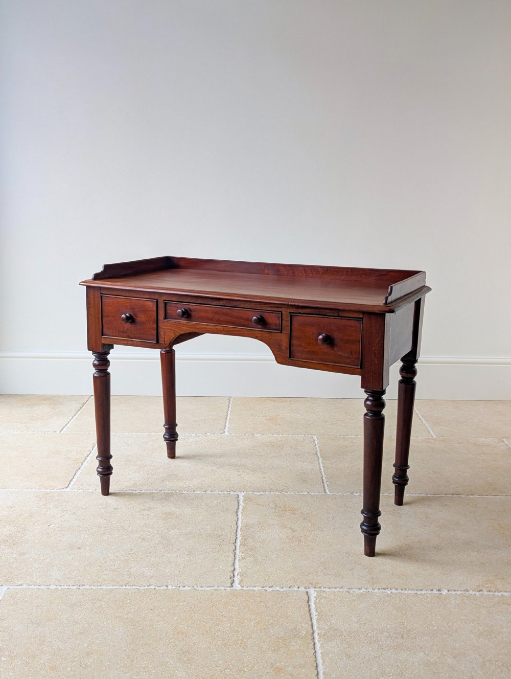 A late Victorian mahogany writing table featuring a low gallery top, three frieze drawers with turned wooden knobs, an arched kneehole, and four turned legs.