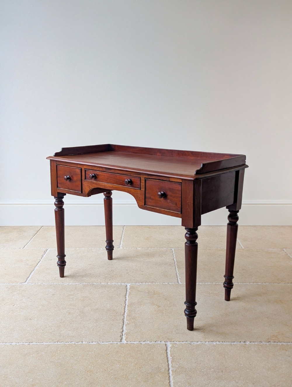 A late Victorian mahogany writing table featuring a low gallery top, three frieze drawers with turned wooden knobs, an arched kneehole, and four turned legs. View VI