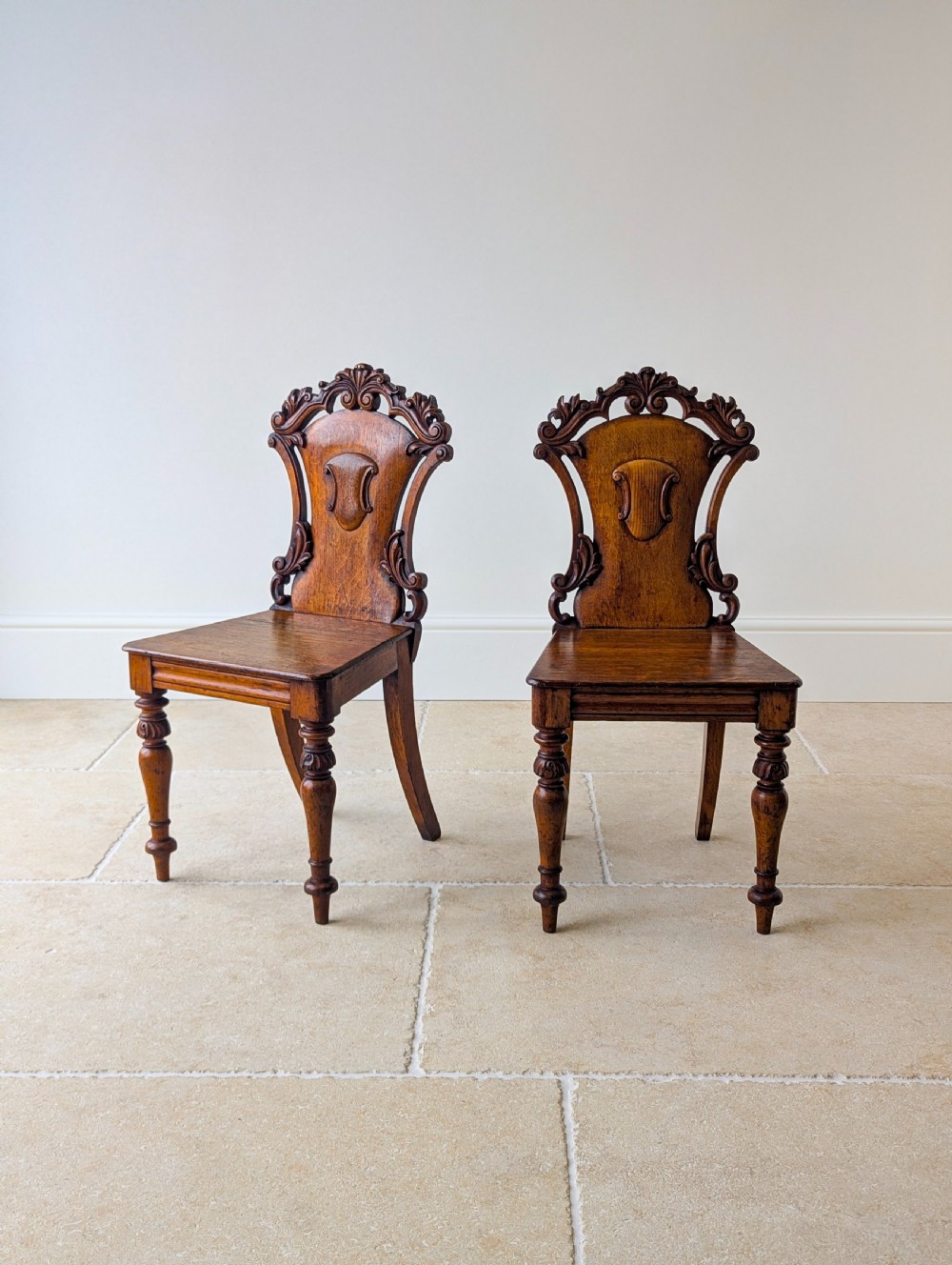 A pair of Victorian oak hall chairs featuring carved cartouche backs, flat wooden seats, and turned front legs.