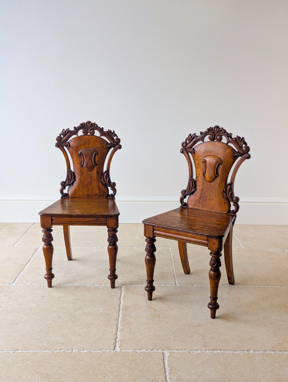 A pair of Victorian oak hall chairs featuring carved cartouche backs, flat wooden seats, and turned front legs. View V