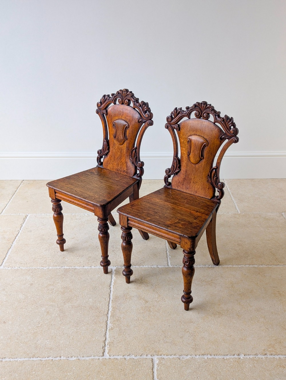 A pair of Victorian oak hall chairs featuring carved cartouche backs, flat wooden seats, and turned front legs. View VII