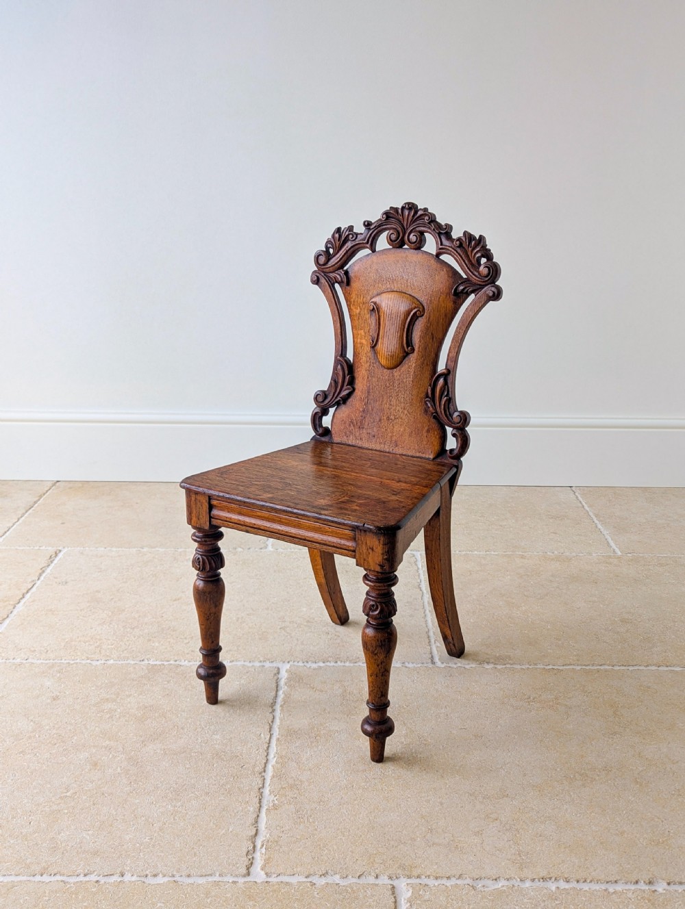 A pair of Victorian oak hall chairs featuring carved cartouche backs, flat wooden seats, and turned front legs. View X
